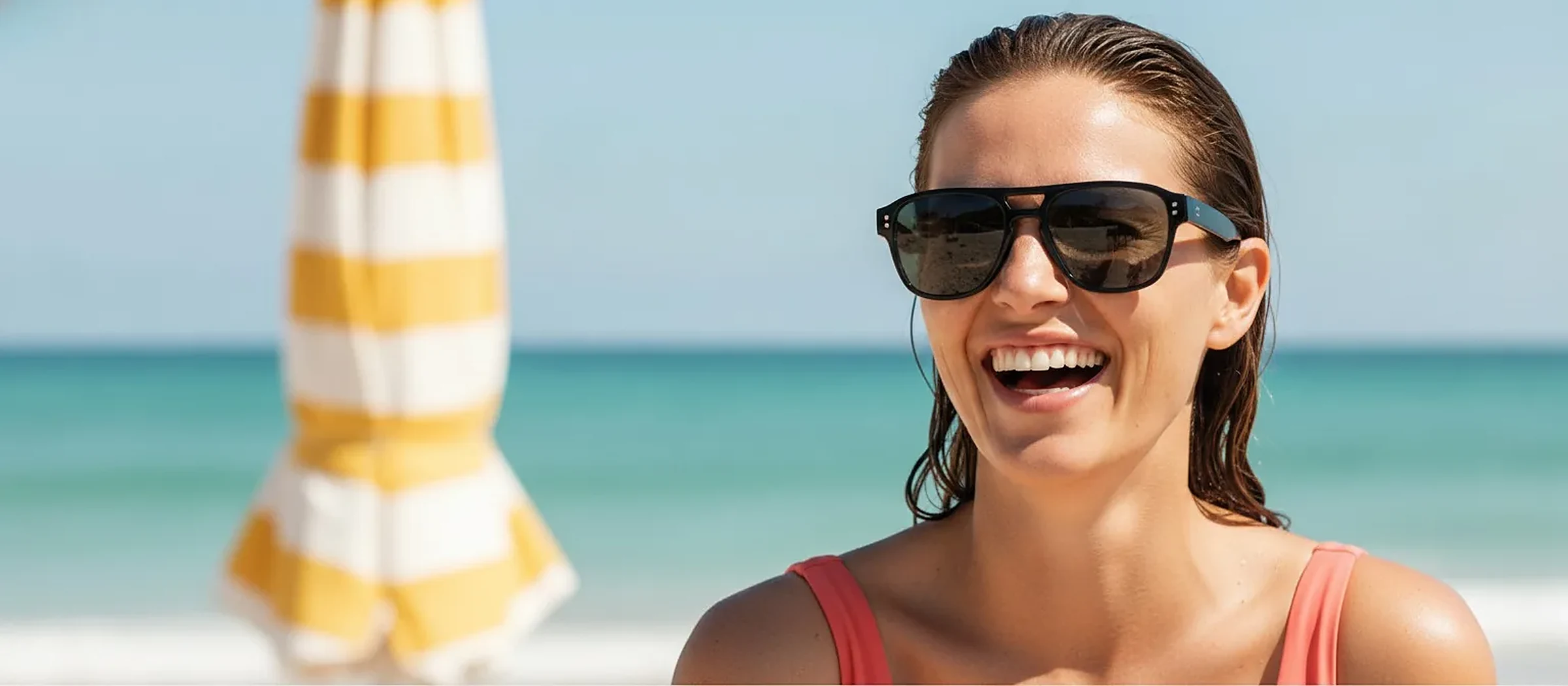 Femme riant portant des lunettes de soleil, à la plage, parasol rayé jaune